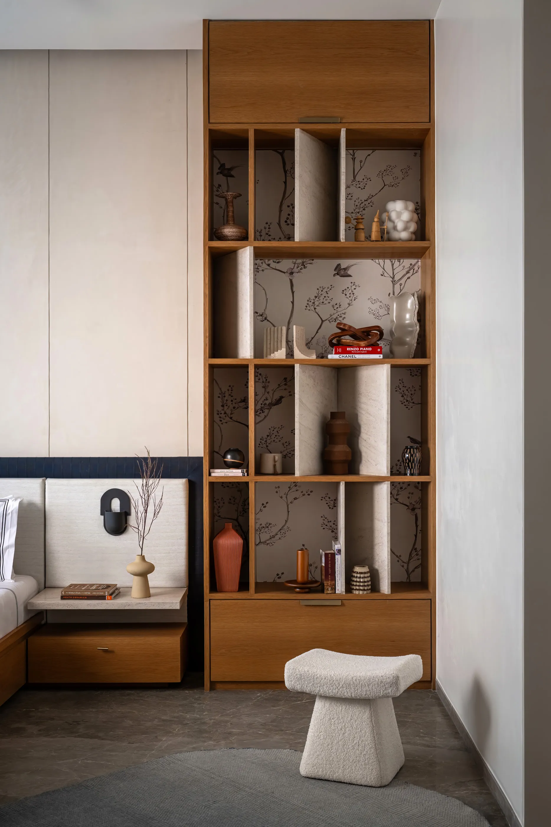 Tall oak shelving with stone dividers and bird-print wallpaper beside a bed, with a boucle stool